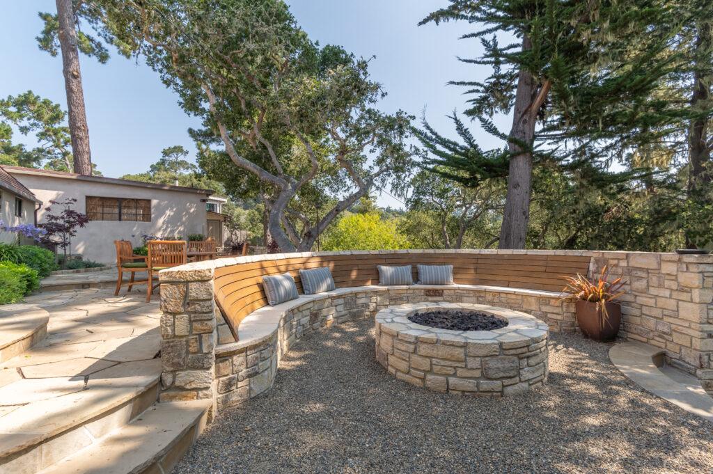 Curved stone and wood bench surrounding a fire pit at the Castle House outdoor renovation in Carmel-by-the-Sea, with oak trees overhead